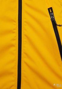 Chaqueta amarilla con una textura suave, presenta detalles de cremallera y costuras en negro. Incluye dos bolsillos con cremallera en la parte frontal.