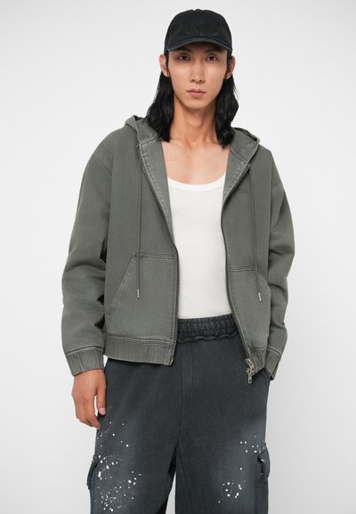 Zelená kapucňová bunda so zipsom a prednými vreckami, skombinovaná so šedými nohavicami s detailmi bielej farby a čiernou čiapkou.