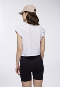 Maglietta bianca cropped con maniche corte, indossata con pantaloni corti neri aderenti; persona con un cappellino beige, vista da dietro.