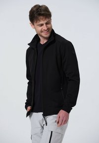 Schwarze Zip-Jacke mit Stehkragen, glatter Textur und seitlichen Taschen. Kombiniert mit einem schwarzen Shirt und hellgrauen Hosen mit Reißverschlüssen.