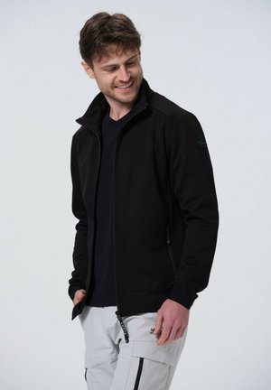 Giacca nera con zip e colletto alto, texture liscia e tasche laterali. Abbinata a una maglietta nera e pantaloni grigio chiaro con cerniere.