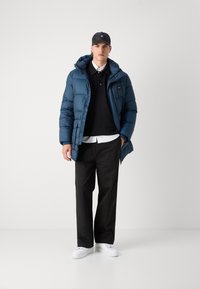 Giacca blu imbottita con cappuccio, dotata di una cerniera frontale e di un logo sul petto, indossata sopra un polo nero e una camicia bianca con pantaloni neri e scarpe bianche.