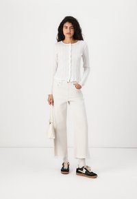Vit ribbad långärmad cardigan med knappar, kombinerad med vida vita byxor och svarta sneakers, hållande en liten vit handväska.