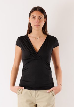 Donna con capelli castani lunghi che indossa una maglia nera con scollo a V e pantaloni beige, in piedi con le mani nelle tasche davanti a uno sfondo semplice.