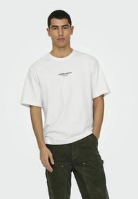Weißes T-Shirt mit kurzen Ärmeln und schwarzem Text "CURATED STAPLES" auf der Brust. Der Stoff wirkt glatt und leicht.