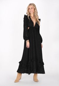 Rochie maxi neagră cu mâneci lungi tip puff, dantelă și tăieturi pe laterale. Prezintă un tiv ondulat și un material texturat. Modelul poartă cizme bej până la glezne.