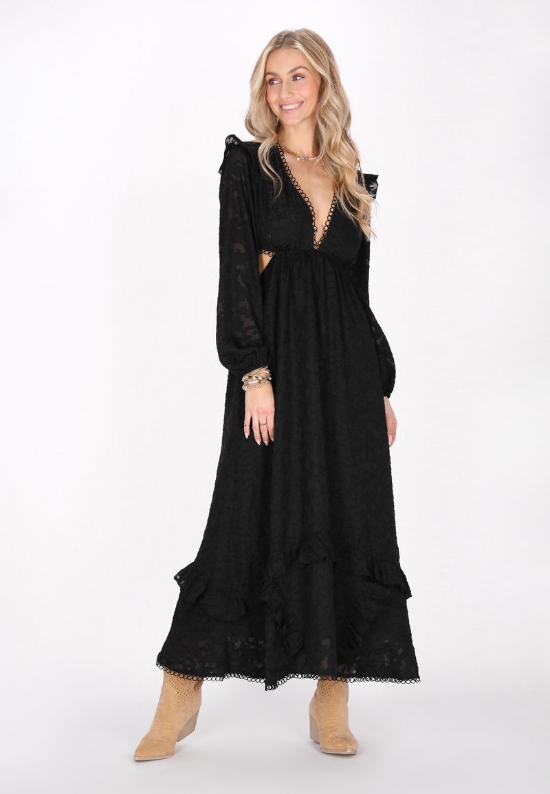 Rochie maxi neagră cu mâneci lungi tip puff, dantelă și tăieturi pe laterale. Prezintă un tiv ondulat și un material texturat. Modelul poartă cizme bej până la glezne.