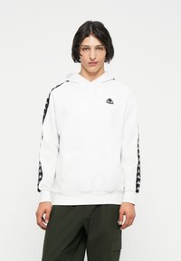 Sudadera blanca con un bolsillo frontal, que presenta un logo negro en el pecho y adornos negros estampados a lo largo de las mangas. Tela de algodón suave.