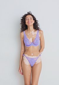 Glunderende vrouw met krullend haar, gekleed in lila kanten lingerie, staand tegen een effen lichtgrijze achtergrond.
