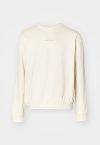 Cream-colored sweatshirt with long sleeves, round neckline, ribbed cuffs, and hem. Features minimal text logo on the chest in a subtle tone.