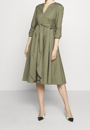 Oliivinvihreä mekko, jossa on V-aukko, vyötäröltä kiristettävä malli ja liehuva helma. Mekossa on pitkät hihat ja vyötäröllä solmittava yksityiskohta. Yhdistetty vaaleanvärisiin korkokenkiin.