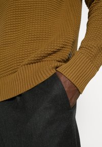 Main à la peau foncée dans la poche d'un pantalon noir, portant un pull en maille texturée jaune moutarde avec des poignets et un ourlet côtelés.