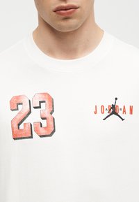 Κοντινό πλάνο ενός λευκού πουκαμίσου με κόκκινο αριθμό 23 και μαύρο λογότυπο Jumpman με κείμενο "JORDAN" στο στήθος.