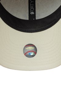 Beige Baseballkappe mit schwarzem Schirm, versehen mit einem glänzenden runden MLB-Aufkleber auf dem Schirm und strukturiertem Stoff auf der Innenseite.