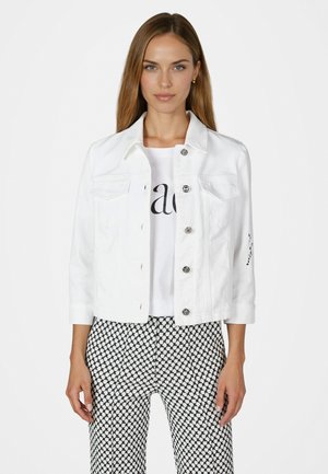Woman wearing a white buttoned jacket over a white printed shirt and black-and-white patterned pants, standing against a plain background.