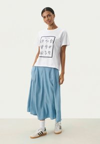T-shirt bianco in cotone con un design floreale a linea, abbinato a una gonna di denim blu. Sneakers con tre strisce nere e calzini bianchi.