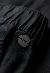 Zwarte stof met een gematteerde afwerking, voorzien van een elastische tailleband en een ronde, zwarte logotag met de tekst "SUPERDRY."