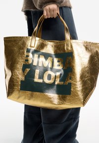 Złota metaliczna torba na ramię z zielonym logo "BIMBA Y LOLA" wyraźnie widocznym. Materiał ma błyszczące, teksturowane wykończenie oraz mocne rączki.