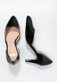 Chaussures à talons aiguilles en cuir verni noir avec un bout pointu, des côtés ouverts et une semelle intérieure texturée. Talon aiguille d'environ 10 cm de hauteur.