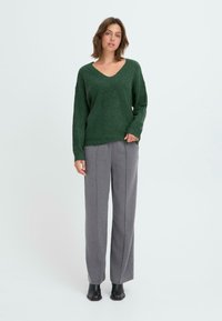 Maglione verde lavorato a maglia con scollatura a V e spalle basse, abbinato a pantaloni grigi chiari a gamba larga e stivaletti neri alla caviglia.