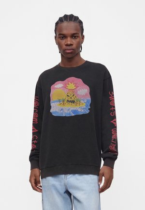 Jeune homme portant un sweat-shirt noir avec un motif coloré de volcan et d'île sur le devant et un texte rouge sur les deux manches, associé à un jean bleu clair.
