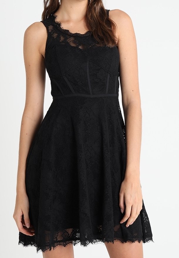 Robe noire en dentelle sans manches avec corsage ajusté, jupe évasée et encolure festonnée portée par une femme sur fond uni.
