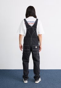 Svarte denim-overaller med justerbare stropper, lomme på brystet og to sidelommer, med hvite sømmer og en logomerking.