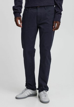 Mężczyzna w granatowych casualowych spodniach, ciemnym swetrze i jasnoszarych niskich trampkach, stojący z rękami luźno opuszczonymi wzdłuż ciała.