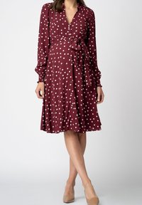Burgundowa sukienka w groszki z długimi rękawami, zapięciem na guziki z przodu, paskiem w talii oraz rozkloszowanym dołem. Materiał ma miękką, gładką teksturę.