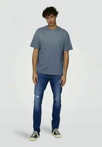 T-shirt gris à manches courtes avec un col rond, associé à un jean slim déchiré bleu et des baskets noires. Ensemble simple et décontracté.