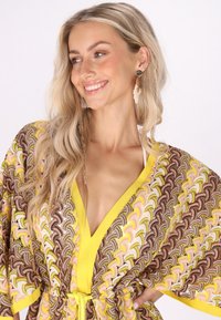 Šaren kaftan sa valovitim uzorkom u ružičastoj, žutoj i smeđoj boji, s dubokim V-izrezom, rukavima nalik krilima i detaljem uvezivanja u struku.