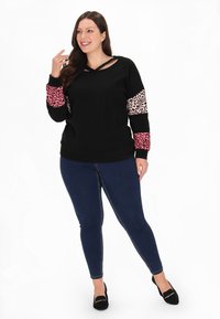 Schwarzer Pullover mit V-Ausschnitt und Ausschnitt-Detail. Lange Ärmel mit rosa und Leoprint-Akzenten. Kombiniert mit dunkelblauen Jeans und schwarzen Schuhen.