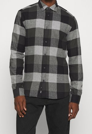 Man wearing a buttoned black and gray checkered long-sleeve shirt with black pants, standing against a plain light background.