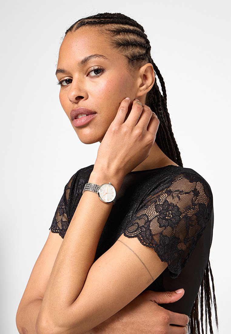 Vrouw met gevlochten haar, gekleed in een zwart kanttop en zilveren polshorloge, die haar gezicht aanraakt met een neutrale uitdrukking tegen een effen achtergrond.