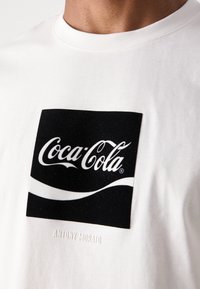 Witte katoenen T-shirt met een groot zwart rechthoekig Coca-Cola logo en een soepele witte golfgrafiek. Bevat "ANTONY MORATO" in reliëftekst.