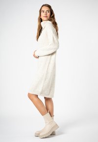 Strickkleid in Creme mit geripptem Muster, ausgestattet mit einem hohen Kragen und langen Ärmeln. Kombiniert mit beige-farbenen Stiefeletten mit dicker Sohle.