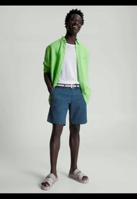 Πράσινο πουκάμισο με κουμπιά, λευκό μπλουζάκι, μπλε σορτς και γκρι σανδάλια. Το σύνολο έχει μια ζώνη με σχέδιο και χαλαρή εφαρμογή.