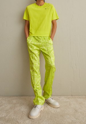 Person iført en knaldrød limegrøn T-shirt og skinnende mønstrede bukser med hvide chunky sko, stående foran en ensfarvet beige væg.