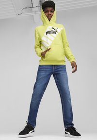 Sudadera amarilla con capucha y diseño del logo de Puma en blanco, bolsillo frontal y vaqueros. El calzado consiste en zapatillas negras con suelas blancas.