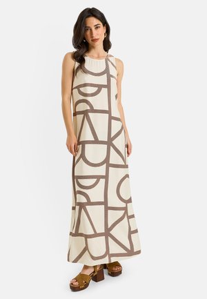 Femme aux cheveux foncés porte une robe longue crème sans manches avec de grands motifs abstraits marron et des sandales compensées marron.