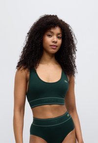Femme aux cheveux bouclés portant un brassière de sport côtelée vert foncé et un slip taille haute, tous deux avec le logo Lacoste sur les bandes élastiques.