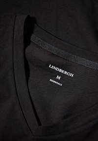 Schwarzes T-Shirt mit V-Ausschnitt, aus glattem Baumwollstoff. Mit einem Etikett versehen, auf dem "LINDBERGH M ESSENTIALS" in Weiß gedruckt ist.