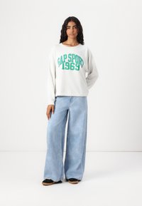 Felpa grigia chiara a maniche lunghe con testo verde "GAP SPORT 1969", abbinata a jeans azzurri a gamba larga e scarpe con plateau nere.