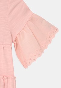 Jasnoróżowy bawełniany top z rozkloszowanym rękawem wykończonym koronką i falbaniastymi brzegami, podkreślający delikatną fakturę i swobodny styl.