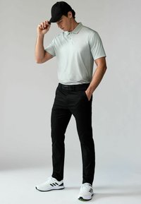 Hellgraues kurzärmliges Polohemd, schwarze Hose und weiße Sportschuhe. Mann mit schwarzem Hut, Hände in den Taschen. Einfaches, modernes Design.