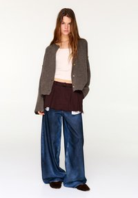 Oversized bruine cardigan, licht tanktopje, gelaagde kastanjebruine rok, losse spijkerbroek en bruine pluizige pantoffels; een casual gelaagde outfit.