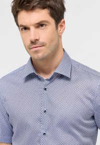 Camicia a maniche corte con bottoni, caratterizzata da un motivo geometrico blu e bianco, con colletto standard e bottoni scuri. Tessuto dalla superficie liscia.