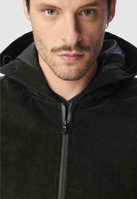 Close-up van een man met lichte stoppels, gekleed in een zwarte rits-hoodie met een opstaande kraag en een kleine zilveren ringoorbel.
