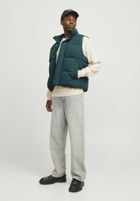 Teal gewatteerde vest met hoge kraag, drukknoopsluiting en voorkruiszakken, gedragen over een lichte trui, gecombineerd met lichte jeans en zwarte schoenen.