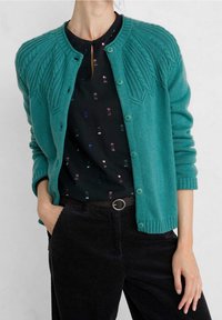 Miętowy dziergany kardigan z teksturowanym karczkiem, okrągłym dekoltem i zapięciem na guziki. W połączeniu z wzorzystą czarną bluzką i sztruksowymi spodniami.